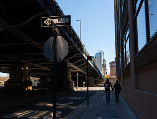 Two 'One Way' street signs intersecting under a bridge in dramatic light with silhouettes of pedestrians in the background
