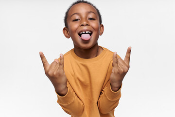 Poster - Close up image of cool handsome African American schoolboy in yellow sweatshirt acting naughty sticking out his tongue, teasing you, showing devil horns gesture, having fun, Heavy metal and hard rock