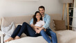 © fizkes - Portrait of young wife and husband hugging, sitting on couch, smiling attractive woman and man cuddling, looking at camera, posing for family photo at home, relaxing on sofa, enjoying lazy weekend