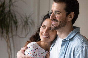 Wall Mural - Close up dreamy young couple cuddling, looking to aside, smiling attractive woman and man wearing glasses hugging, wife and husband visualizing good future, planning, dreaming and thinking