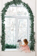 © Olga Mishyna - Little girl weared in pink holiday dress sitting on an arch window in white living room decorated for Christmas celebration with spruce garland. New year eve, snowy weather. Happy Holidays