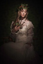 This Image Is A Stylized Painterly Portrait Of Young Woman In Fancy White Lace Antique Dress With A Holly Crown On , Loving Holding An Old Brass Statue And Looking Downwards.