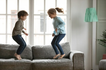 Energetic little kids siblings jumping barefoot on couch, enjoying spending free weekend leisure time together at home. Happy small preschool brother and sister involved in funny domestic activity.