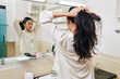© DragonImages - Pretty young Chienese woman in white shirt making high ponytail in front of mirror in bathroom