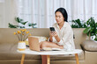 © DragonImages - Pretty young Asian businesswoman in pajamas sitting of sofa after working on laptop and checking messages from colleague in smartphone