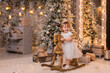 © Юлия Немченко - Selective focus. Close-up portrait of a little girl riding a horse on a wooden vintage rocking chair against the background of a Christmas tree. Christmas and New Year. Holiday concept.