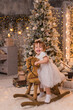 © Юлия Немченко - Selective focus. Close-up portrait of a little girl riding a horse on a wooden vintage rocking chair against the background of a Christmas tree. Christmas and New Year. Holiday concept.
