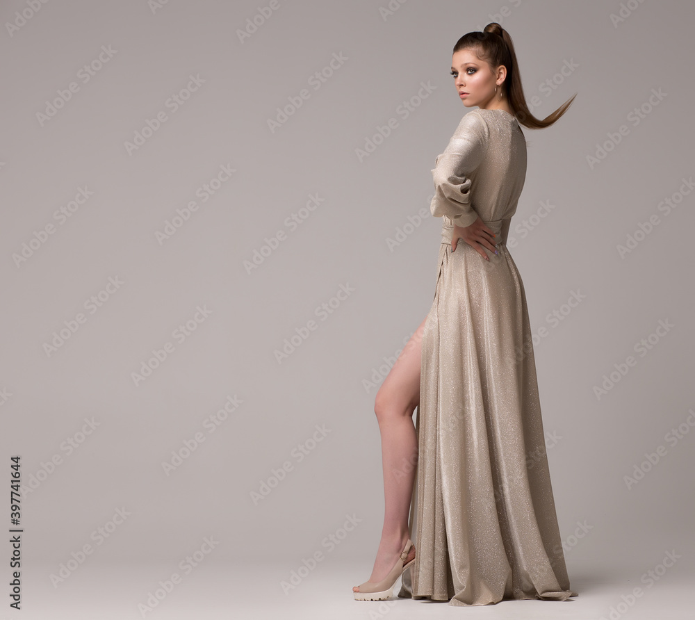 Photo Stock A woman in a long dress poses in the studio in full growth. She stands half a turn, hand at the waist, leg extended forward. Long light dress. | Adobe Photo Stock A woman in a long dress poses in the studio in full growth. She stands half a turn, hand at the waist, leg extended forward. Long light dress. | Adobe