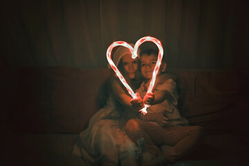 Kids in red hat holding big luminous heart formed with two candy sticks over dark background