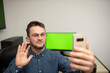 © FarukPhotography - Young caucasian businessman with eyeglasses holding a mobile phone and talking to somebody. Having online meeting. Happy businessman having a good day at job. Computer with green screen in background.