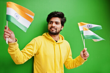 Urban young hipster indian man in a fashionable yellow sweatshirt hold flags of India in hands. Cool south asian guy wear hoodie isolated on green background.