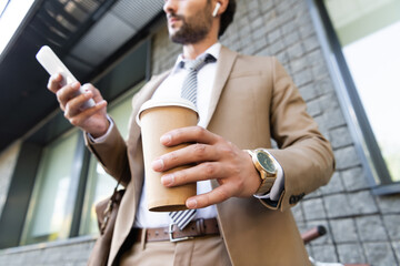 Wall Mural - cropped view of businessman in wireless earphones and suit holding coffee to go and smartphone near building