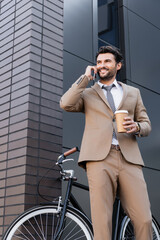 Wall Mural - low angle view of cheerful businessman talking on smartphone and holding paper cup near bicycle and building