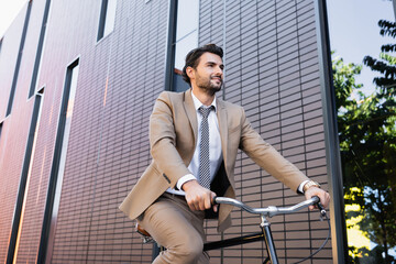 Wall Mural - low angle view of bearded man in suit riding bicycle, smiling and looking away near building