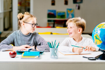 Canvas Print - Smiling schoolgirl talking with friend, looks at each other, smiling while writing in notebook in classroom at desk