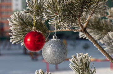 Close up shot of a glittering white and blurred shiny red Christmas balls hanging off a Christmas fir tree outside, all partially covered in snow. Blurred building as a backdrop