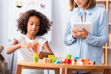 African american girl playing with wooden blocks near psychologist holding digital tablet with blurred coffee table on background