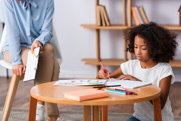 Wall Mural - African american girl drawing with colored pencils on coffee table near psychologist with clipboard on blurred background