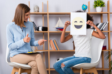 Wall Mural - Smiling psychologist with thumbs up looking at african american girl covering face with happy expression on paper during consultation