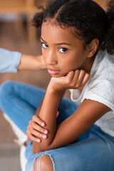 Wall Mural - Upset african american girl with crossed legs looking at camera with blurred hand of psychologist on shoulder on background