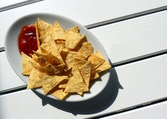 Nachos with red sauce on a white plate under sunlight. White painted slatted wood table on background