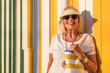 © VISTA by Westend61 - Fashionable senior woman with eyeshield and sunglasses standing in front of colorful striped wall