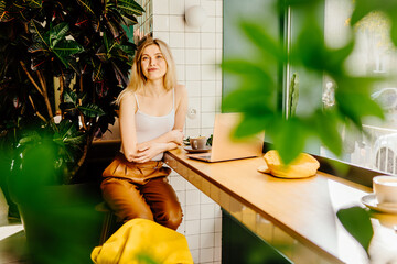 Smiling happy beautiful blond business woman wearing white top relaxing using laptop.Young hipster girl freelancer working and sitting on chair in green plant cafe decorated around her.