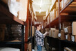 © zzzdim - Short-haired woman taking list of warehouse inventory. Side view. Tall shelves with boxes and tags on both side of her. She wears sheckered shirt and glasses.