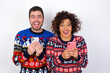 © Roquillo - Excited Young couple wearing Christmas sweater standing against white wall holding smartphone and looking amazed to the camera after receiving good news.