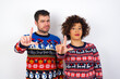© Roquillo - Young couple wearing Christmas sweater standing against white wall making fun of people with fingers on forehead doing loser gesture mocking and insulting.