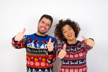 Wall Mural - Young couple wearing Christmas sweater standing against white wall approving doing positive gesture with hand, thumbs up smiling and happy for success. Winner gesture.