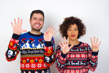 Wall Mural - Young couple wearing Christmas sweater standing against white wall showing and pointing up with fingers number nine while smiling confident and happy.