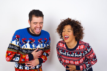 Wall Mural - Young couple wearing Christmas sweater standing against white wall smiling and laughing hard out loud because funny crazy joke with hands on body.