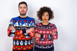 © Roquillo - Serious pensive Young couple wearing Christmas sweater standing against white wall feel like cool confident entrepreneur cross hands.