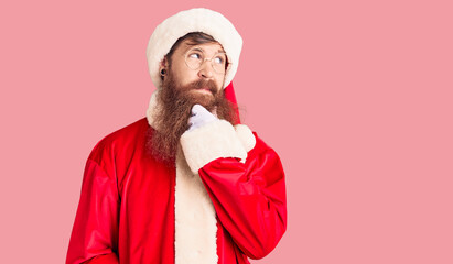 Sticker - Handsome young red head man with long beard wearing santa claus costume with hand on chin thinking about question, pensive expression. smiling with thoughtful face. doubt concept.