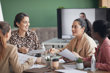 Wall Mural - Multi-ethnic group of businesswomen discussing project during meeting in office with remote participant in background, copy space