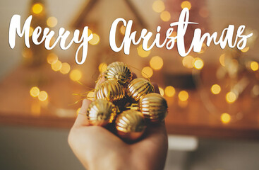 Merry Christmas text handwritten on hand holding modern gold baubles on background blurred illumination lights. Greeting card, modern seasons greetings. Happy holidays