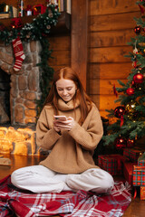 Smiling young redhead woman wearing winter sweater using mobile phone sitting on floor near Christmas tree with bright lighting at cozy living room with festive interior, holiday New Year eve.