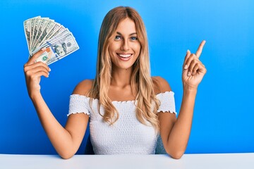 Poster - Beautiful blonde young woman holding dollars smiling happy pointing with hand and finger to the side