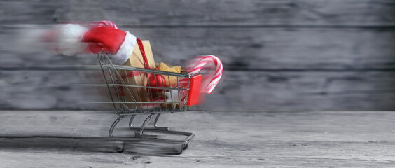 Fast small shopping cart with presents, christmas decoration, sweets and Santa Claus hat is driving on rustic wooden planks for last minute online gifts, panoramic format with copy space, motion blur