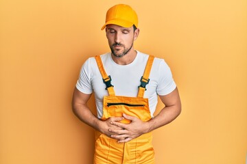 Poster - Young handsome man wearing handyman uniform over yellow background with hand on stomach because indigestion, painful illness feeling unwell. ache concept.