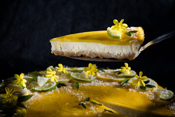 Piece of delicious homemade lemon cheesecake with lime and lemon slices Garnished with yellow flowers and green verbena leaves and coconut flakes. 