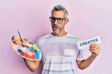 Sticker - Middle age grey-haired man holding palette and creative message paper in shock face, looking skeptical and sarcastic, surprised with open mouth