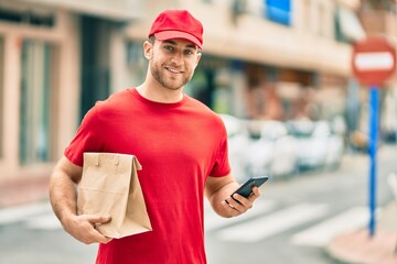 Sticker - Young caucasian deliveryman using smartphone and holding delivery paper bag at the city.