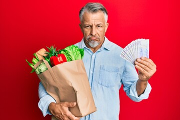 Poster - Middle age grey-haired man holding groceries and swedish krona banknotes depressed and worry for distress, crying angry and afraid. sad expression.