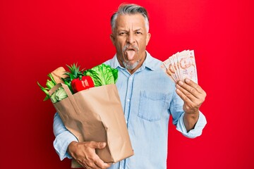 Wall Mural - Middle age grey-haired man holding groceries and singapore dollars banknotes sticking tongue out happy with funny expression.