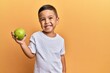 Adorable latin toddler smiling happy holding green apple looking to the camera over isolated yellow background.