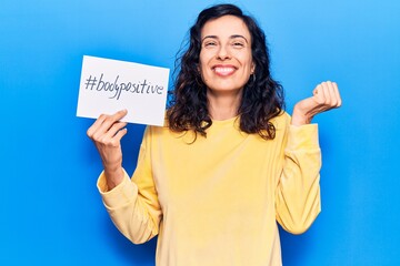 Wall Mural - Young beautiful hispanic woman holding paper with hashtag body positive screaming proud, celebrating victory and success very excited with raised arm