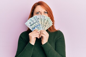 Canvas Print - Beautiful redhead woman holding dollars close to face clueless and confused expression. doubt concept.