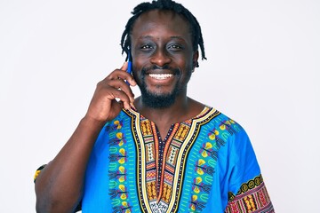 Poster - Young african american man with braids wearing ethnic tshirt having conversation talking on the smartphone looking positive and happy standing and smiling with a confident smile showing teeth